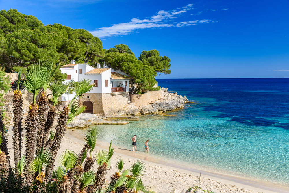 Plaża Cala Gat na Majorce z turkusową wodą, skałami i palmami przy nadmorskiej zabudowie.