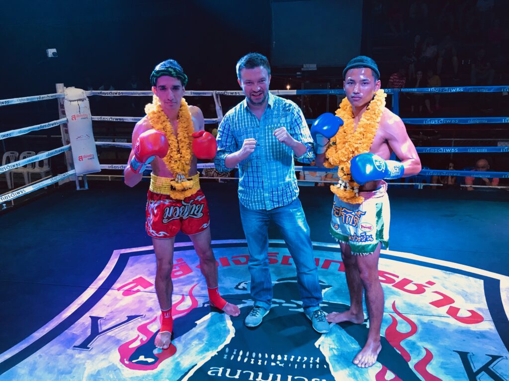 Mężczyzna stojący na ringu pomiędzy dwoma zawodnikami muay thai w rękawicach bokserskich i girlandach z żółtych kwiatów.