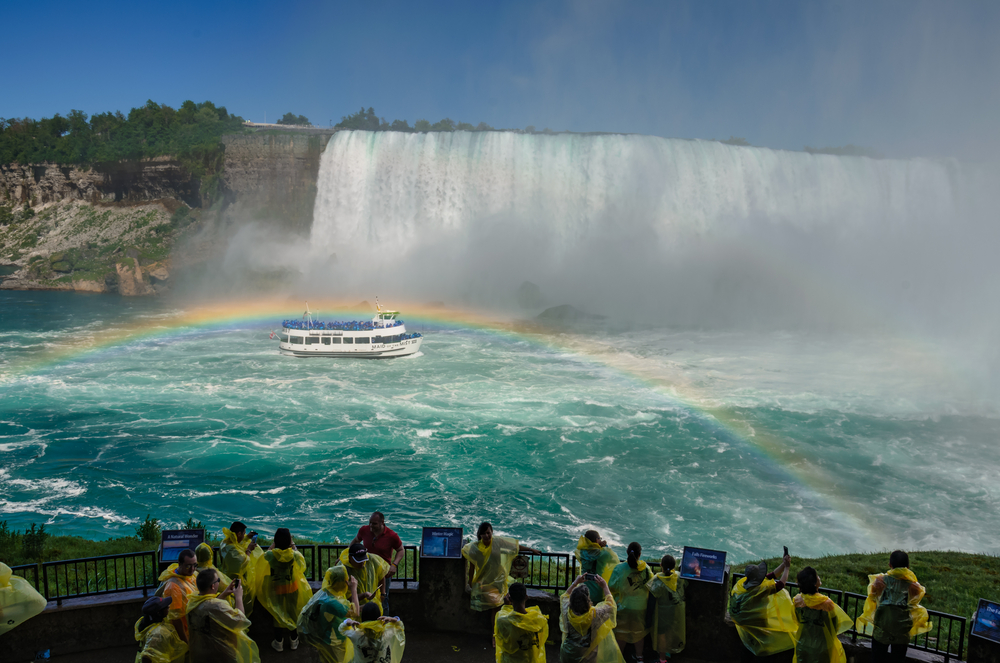 Turyści w żółtych pelerynach na tarasie przy wodospadzie Niagara oraz statek płynący u jego podnóża.