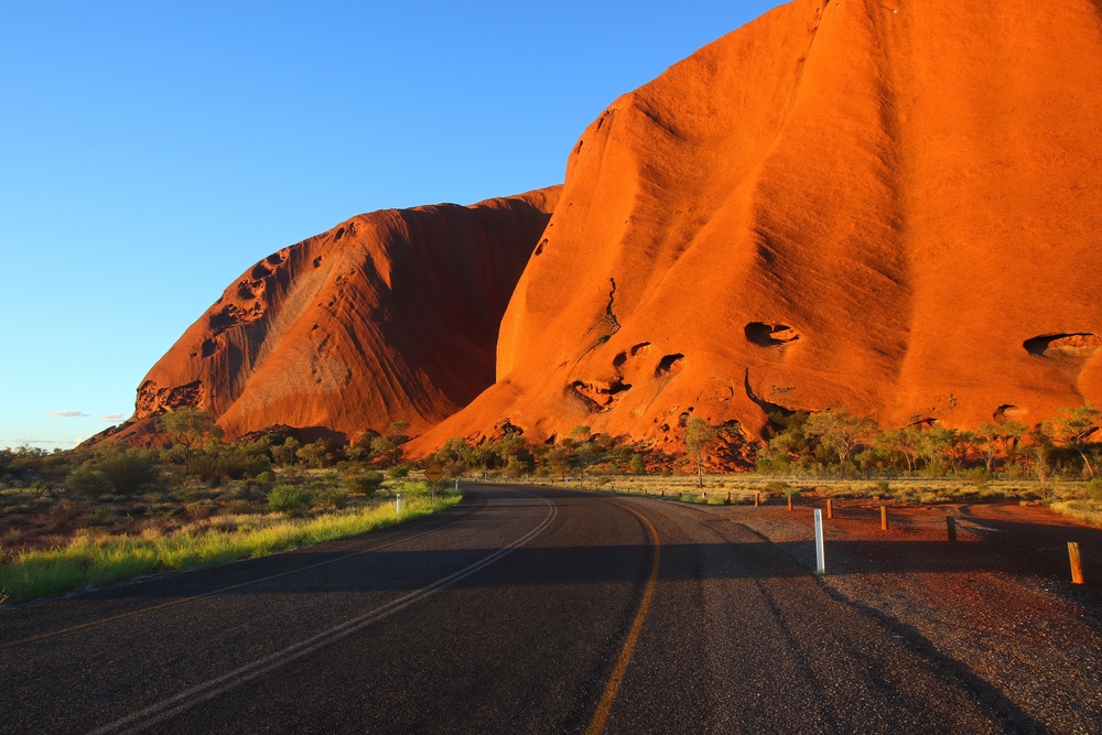 Droga prowadząca wzdłuż czerwonego masywu Uluru w Australii, pod błękitnym niebem.