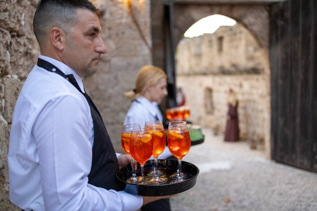 Kelner z tacą kieliszków aperitifu przy kamiennej bramie podczas realizacji TRCO na Rodos.