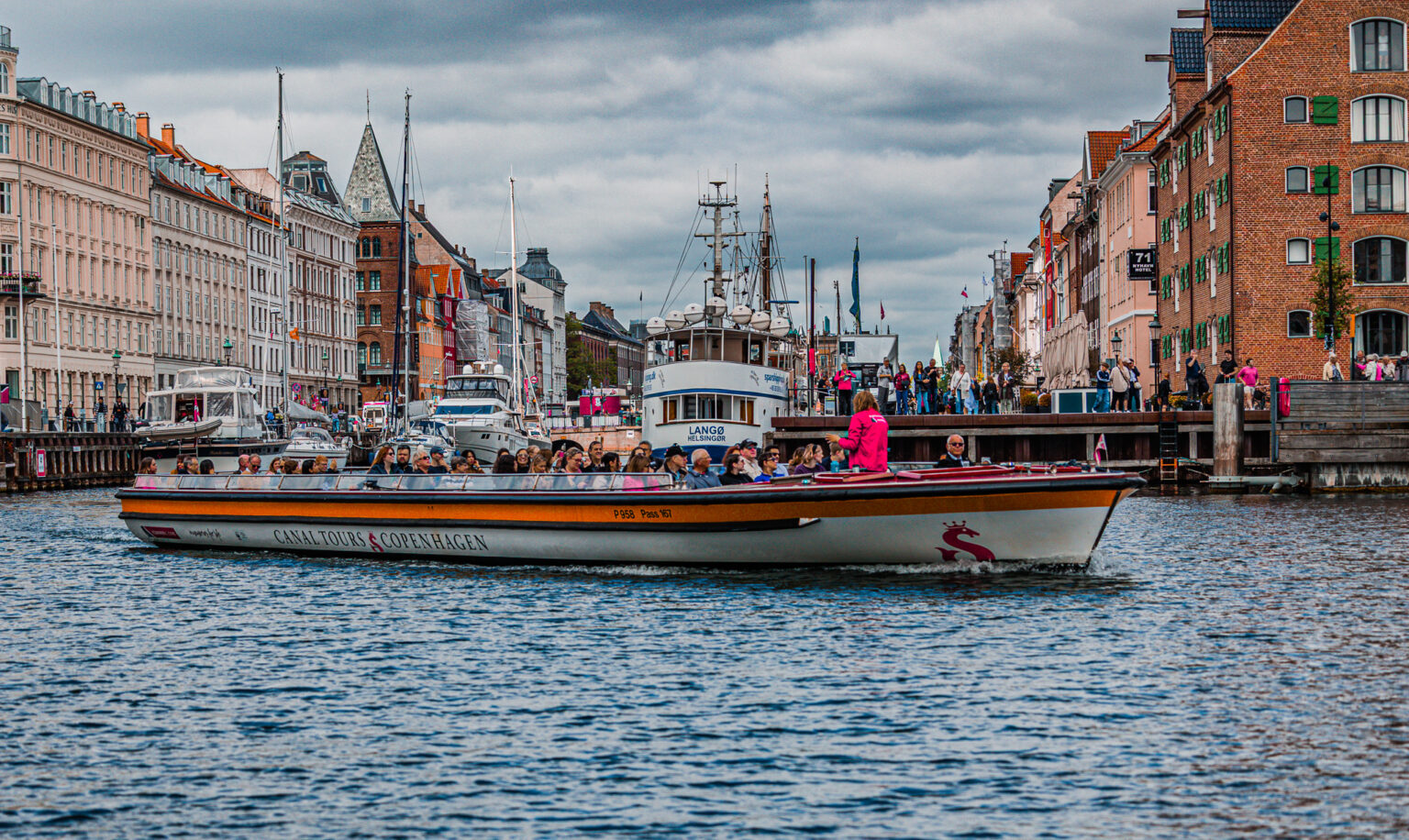 Łódź z pasażerami na kanale Nyhavn w Kopenhadze, na tle kolorowych kamienic i zacumowanych statków.