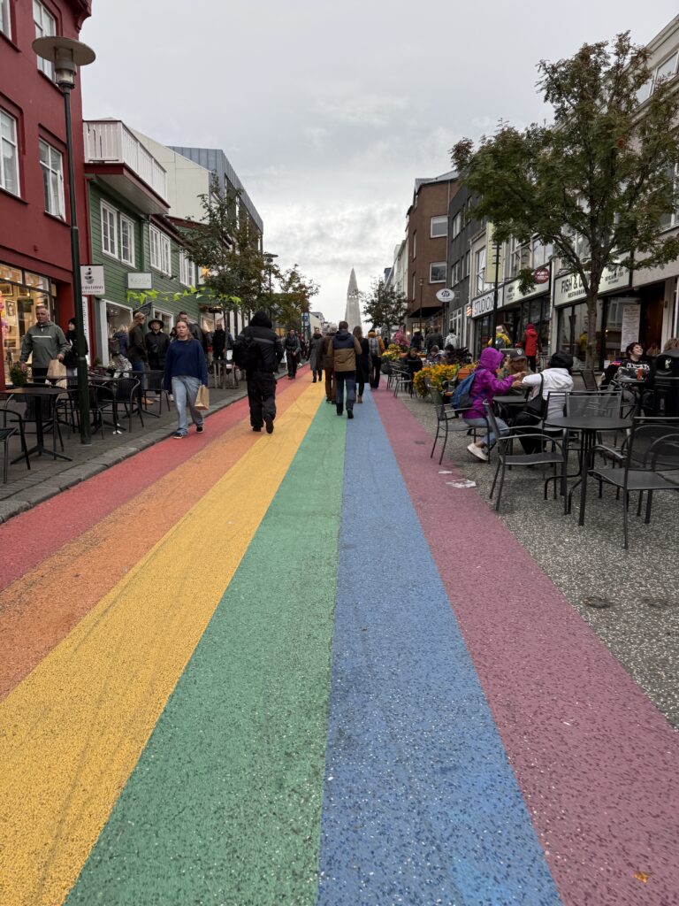 Tęczowa ulica w centrum Reykjavíku, kawiarniane stoliki i kościół Hallgrímskirkja w oddali.